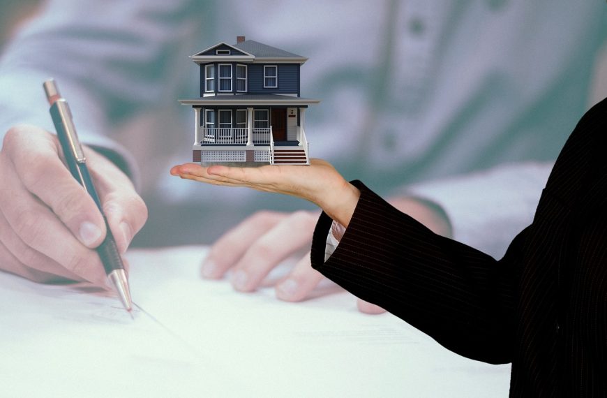 Hand houdt huismodel vast terwijl op de achtergrond een contract wordt ondertekend