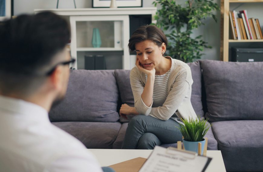 Vrouw in gesprek met adviseur aan tafel in huiselijke omgeving met paarse bank en kamerplanten